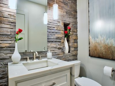 The big-impact look in this powder room results from the contrast of texture, color, and line. The rough-textured, earthy tone stone brings nature inside, while the clean-lined white vanity and countertop provides textural contrast. In addition, the sink, vanity, mirror and art niche provide strong angles, while the lines on the toilet and pendants provide curves.